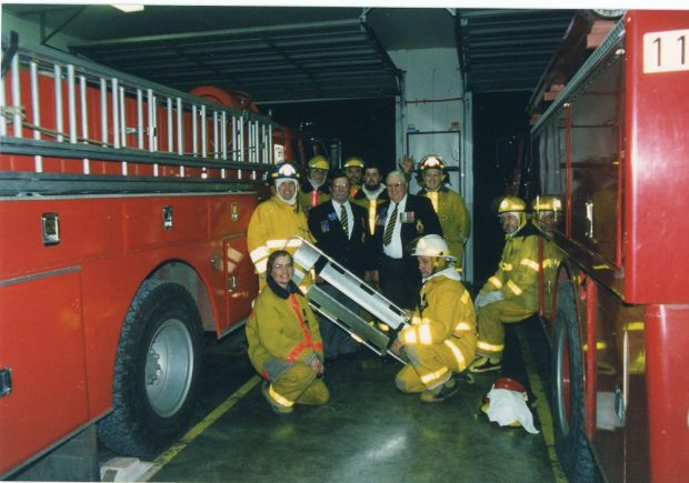 Seven people in firefighting gear and two men in suits with badges between two fire trucks.