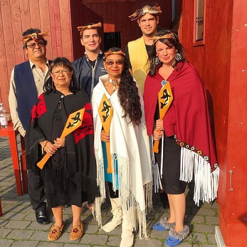 Six people, three men, and three women, standing together dressed in T'Sou-ke First Nations regalia. The three women in the front hold wooden paddles with images of orca whales on them.