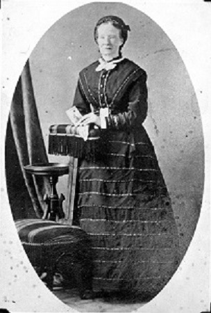 A black and white portrait photo of a woman. She wears a high neck blouse with a white neckerchief and a full, striped skirt. Her hands are resting on a cushioned arm rest, and she holds in her hand what appears to be a card.