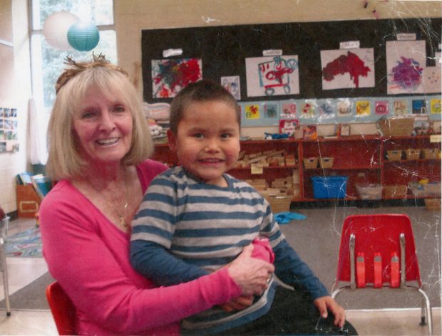 Woman in a classroom holding a young boy in her lap. In the background are children's paintings on the wall, and toys and various education tools in baskets.