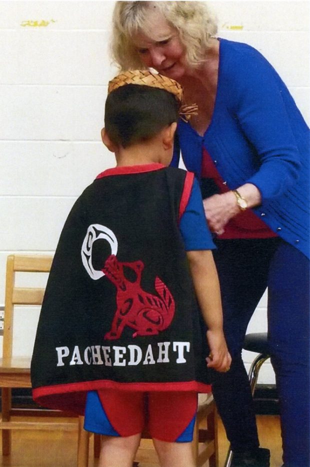 Woman adjusting cape of young boy. Cape features the words Pacheedaht with an image of a wolf and the moon. The design uses traditional Northwest Coast form lines.
