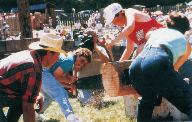 Two women hand bucking a log while a crowd watches. The log is attached with nails to a larger log in a bracket.