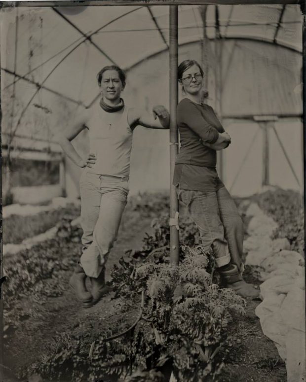 A sepia photograph of two women leaning against a pole in a greenhouse.