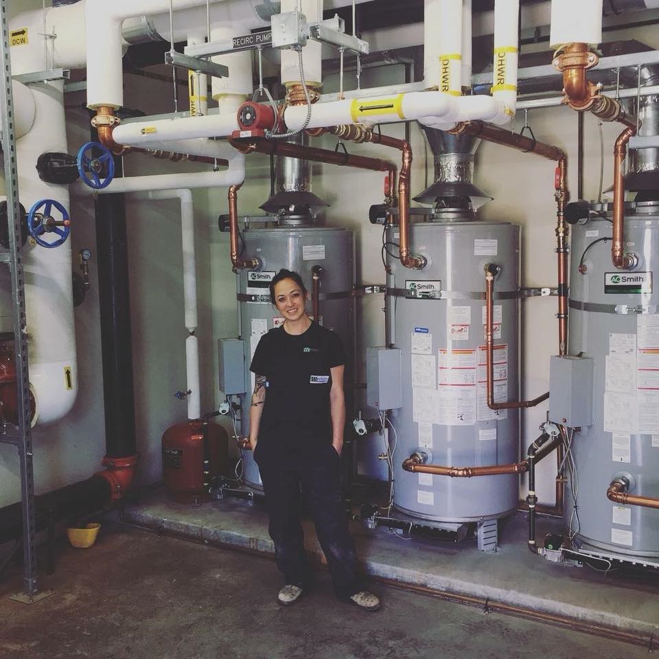 A woman standing in front of three boilers and a network of tubing.