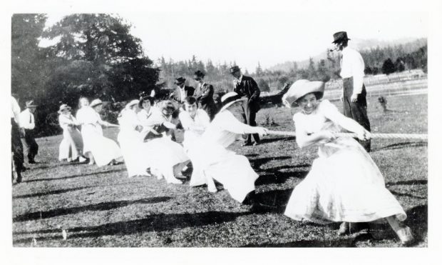 A black and white photograph of several women dressed in dresses and hats pull on a long rope together, smiling.