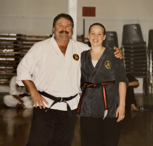 A man and a young woman stand beside each other wearing martial arts uniforms. She wears a black belt with a red stripe and he wears an all-black belt.