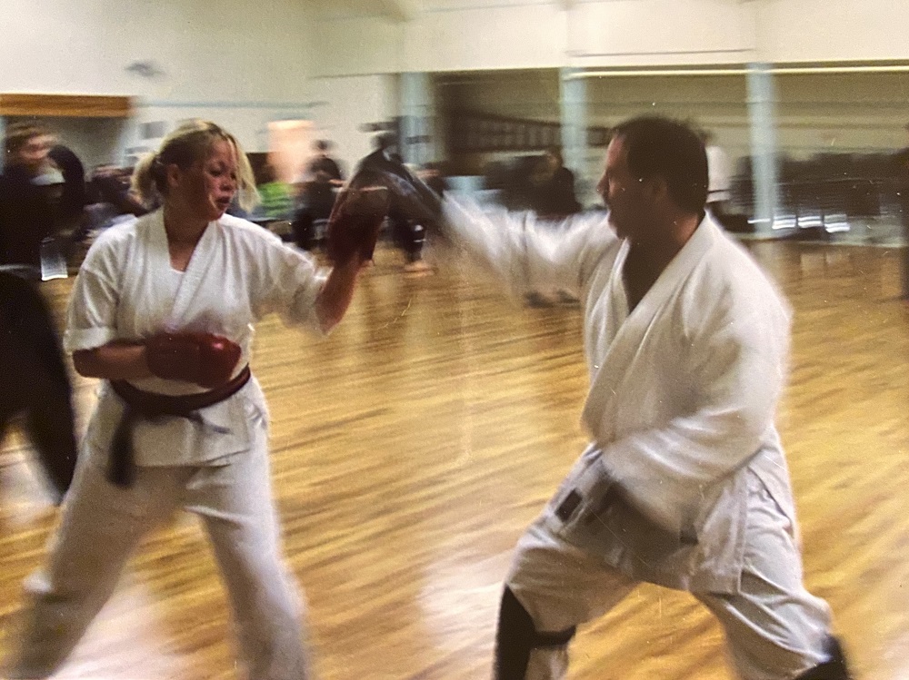A young woman and a man in martial arts uniforms and gear in a gym, in motion, hold their arms up in punching and blocking positions.
