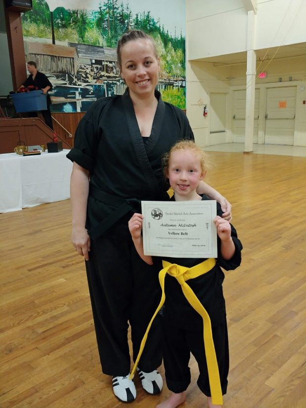 A woman holding the shoulder of her young daughter, who is holding up a certificate for her yellow belt in karate.