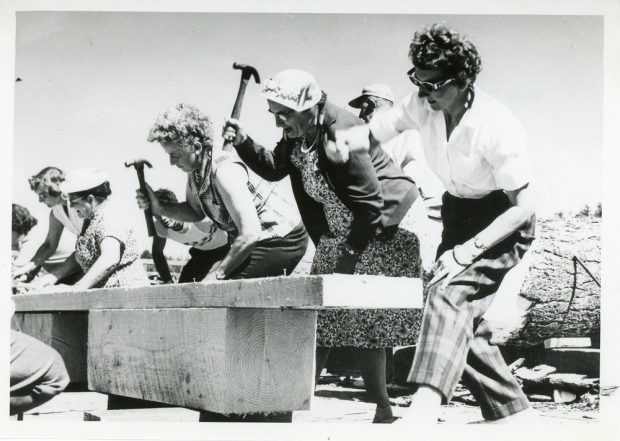 A black and white photograph of a line of women with arms raised holding hammers, hammering nails into a piece of wood.