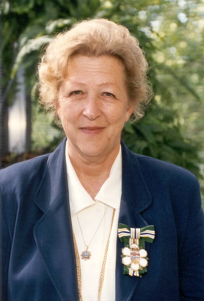 A portrait photo of a woman with a badge on her lapel with an insignia representing a stylized dogwood flower under a bow made of green, gold, white and blue ribbon.