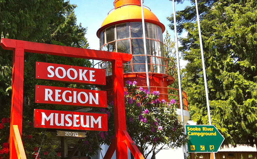 An outdoors scene with a large red wooden sign at the foreground, which reads: "Sooke Region Museum". Behind the sign, towards the center of the picture, we see a large red cylindrical tower - the Triangle Island lighthouse - between the trees.