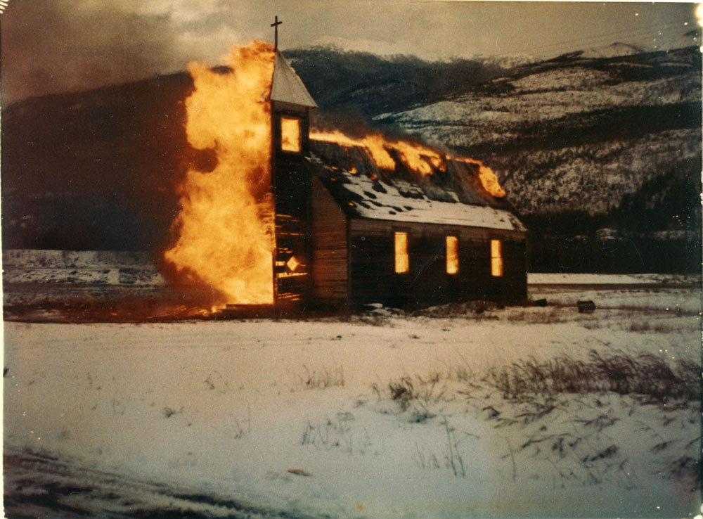 A coloured photograph showing large orange flames burning down a church on a snowy day. Mountains are in the background.