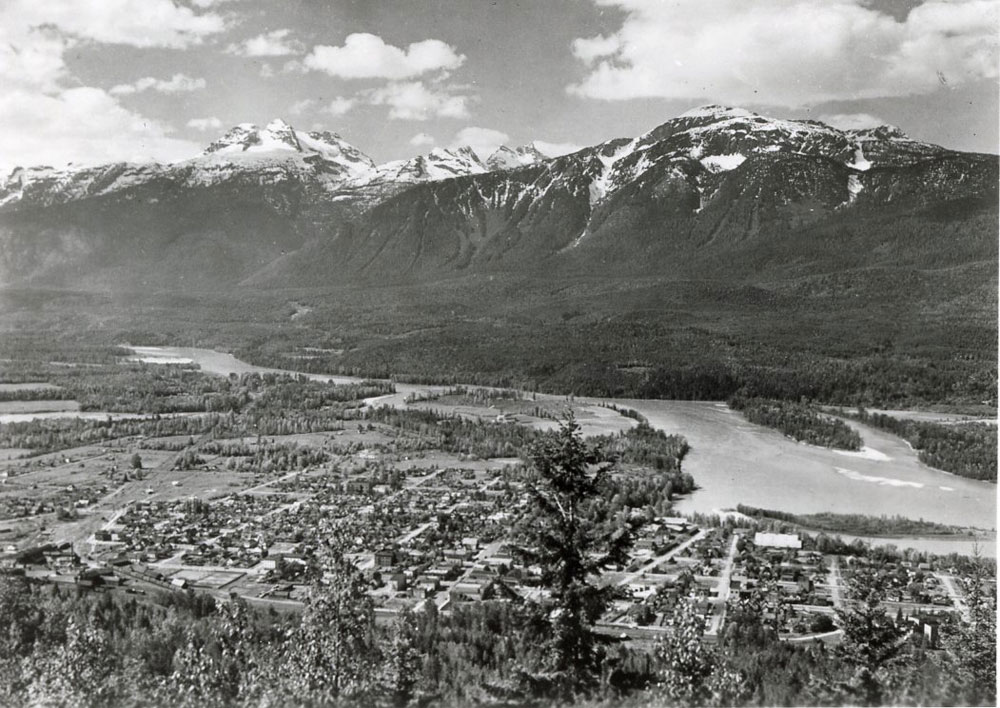 A triple-peaked glacier and other mountains tower over a small city, with a river running between them. The black-and-white photograph was taken from a mountainside, providing a birds-eye view of the city below.