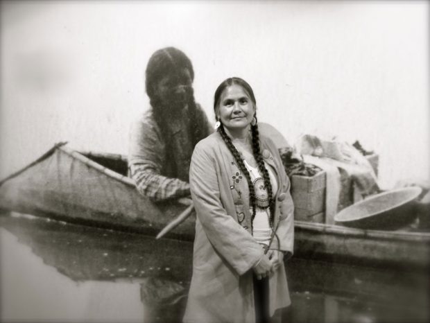 This black-and-white photograph shows an Indigenous woman standing in front of an enlarged archival photograph of an Indigenous woman in a canoe with bows sloping down to the water. Both woman have long black hair, braided.
