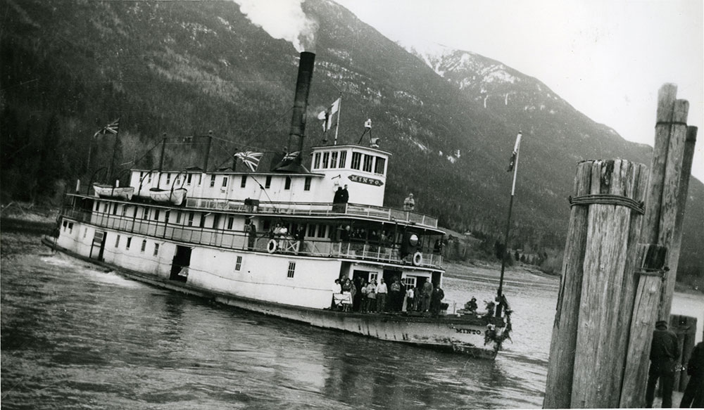 Black-and-white photograph. A steamship on the water with mountains in the background. The steamship has people on board the front, multiple flags sticking out of the ship, and a large wreath on the front of the boat.