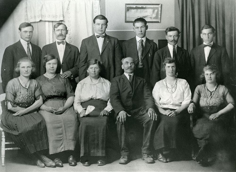 Portrait de famille en noir et blanc de huit personnes en tenue formelle (vestons et jupes). Six hommes sont debout à l’arrière, cinq femmes sont assises à l’avant et un homme est assis au centre. La photographie a été prise dans une pièce avec des rideaux des deux côtés et une petite photo encadrée au centre.