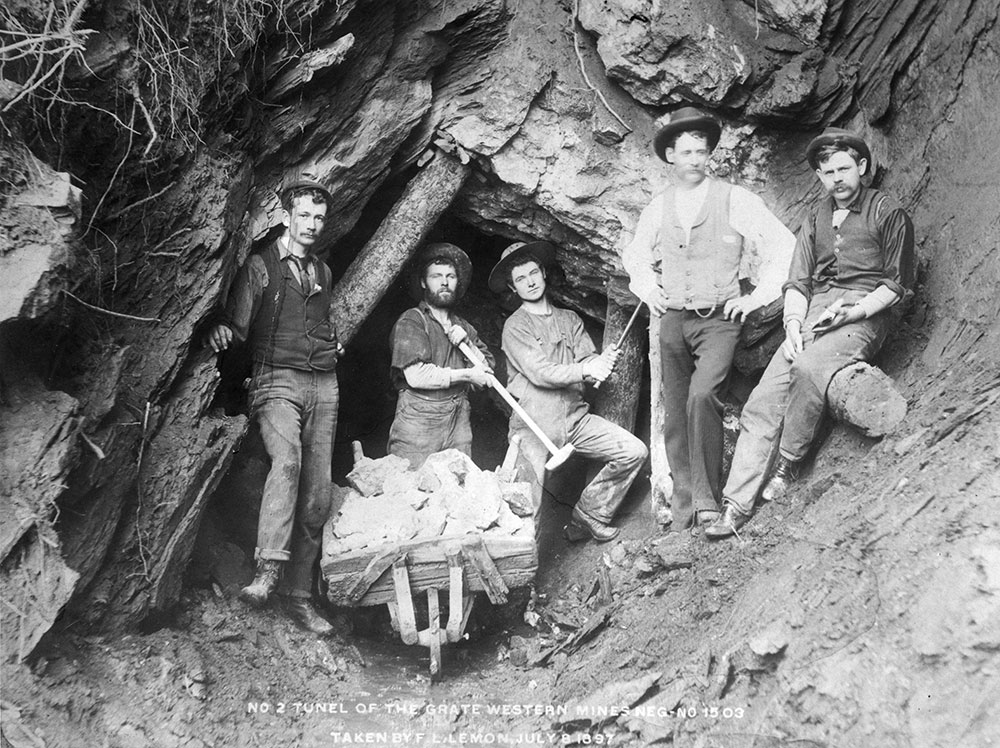 A black-and-white photograph of five miners posing outside of a tunnel. The two miners in front of the tunnel entrance are holding long mining hammers with a wheelbarrow in front of them.