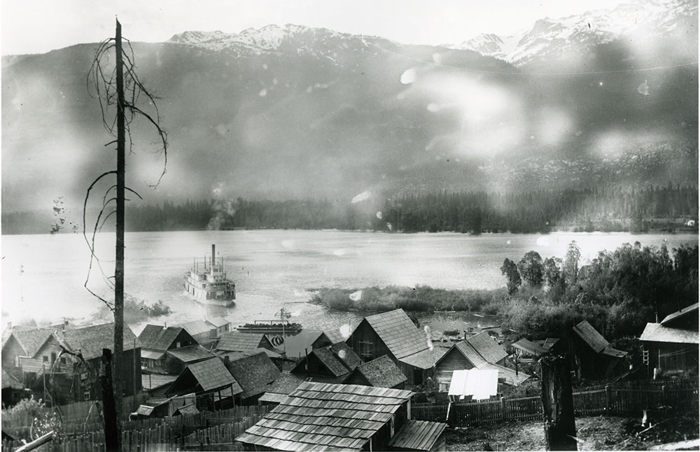 Black-and-white photograph. View of a town, showing multiple houses, angled towards a body of water. A steamship on the water approaches the town. Mountains are in the background. A tall and narrow tree stump with some branches intersects the photo on the left.
