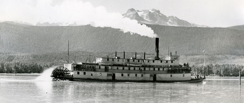 A three-tier paddlewheel steamboat is travelling through the water. In the background is the shoreline and mountain peaks.