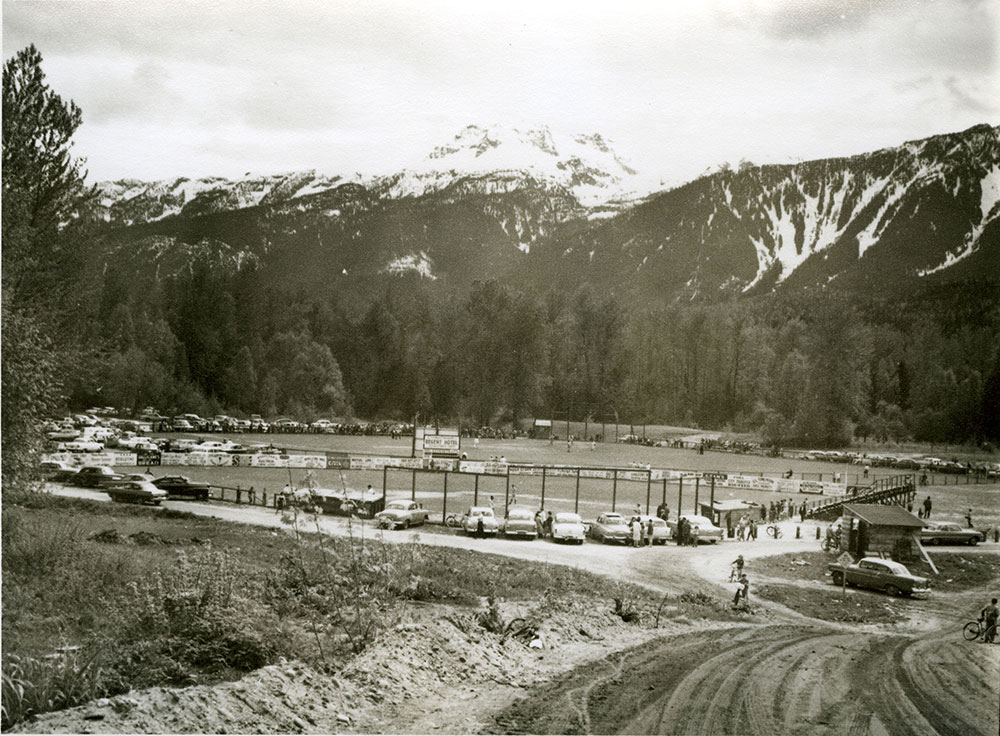 A black-and-white photograph of a baseball field. Cars and people surround the outside of the field. Mountains and trees are in the background. The start of a dirt road is shown on the right before the field.
