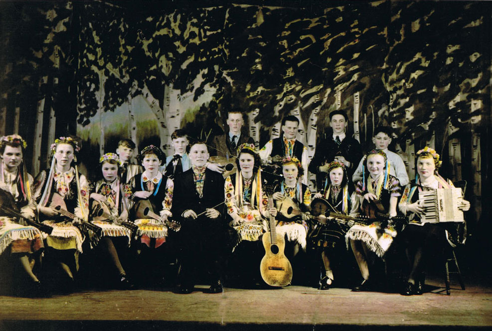 The hand-coloured photo shows fifteen children and one adult gathered on a stage. They are wearing traditional Ukrainian outfits, and each person is holding an instrument.