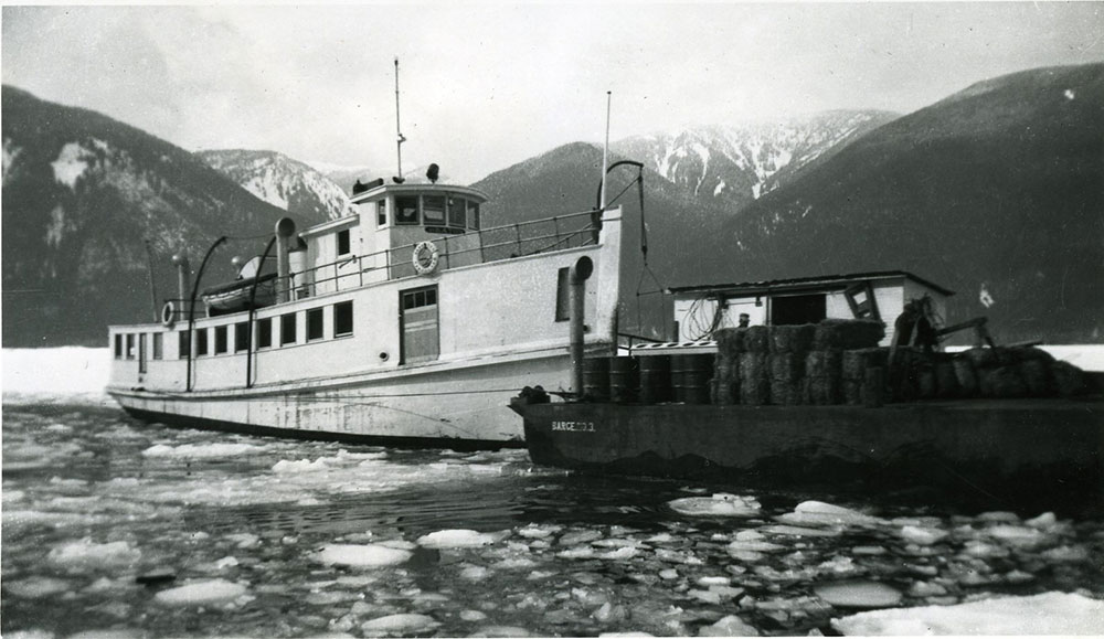 Black-and-white photograph. A small steamship, with a barge attached, sits on icy water. Mountains are in the background.