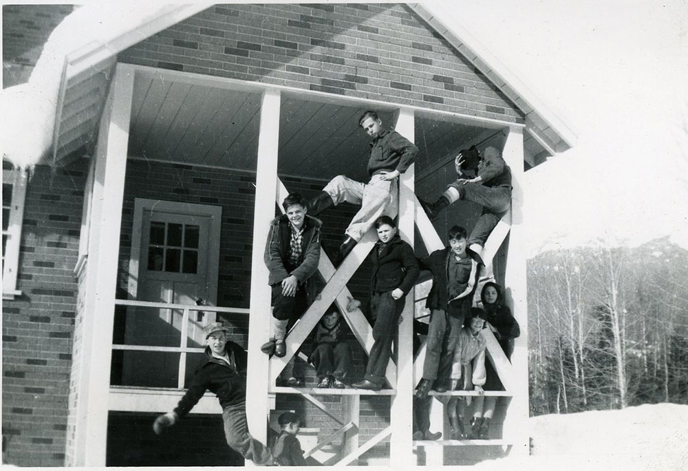 Black-and-white photograph of children posing on the porch of a small brick building. The porch has criss-crossed beams, and the children are posing on top and between the beams.