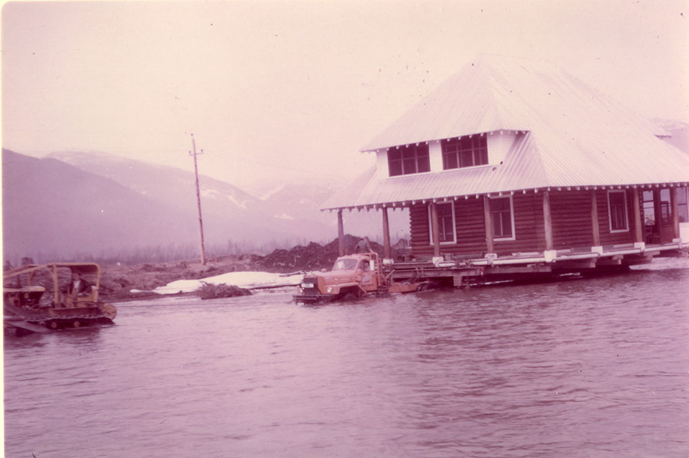 A bulldozer is driving across a river, with a tow rope attached to a large Apex Moving Company truck which is also driving across the river. On the flatbed of the truck is a two-storey log farmhouse. The shore and distant mountains are visible in the background.