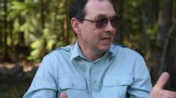Ron Gawiuk - a white man, with black hair, sunglasses, and a blue button down shirt. He is gesturing with his hands. The forest is in the background.