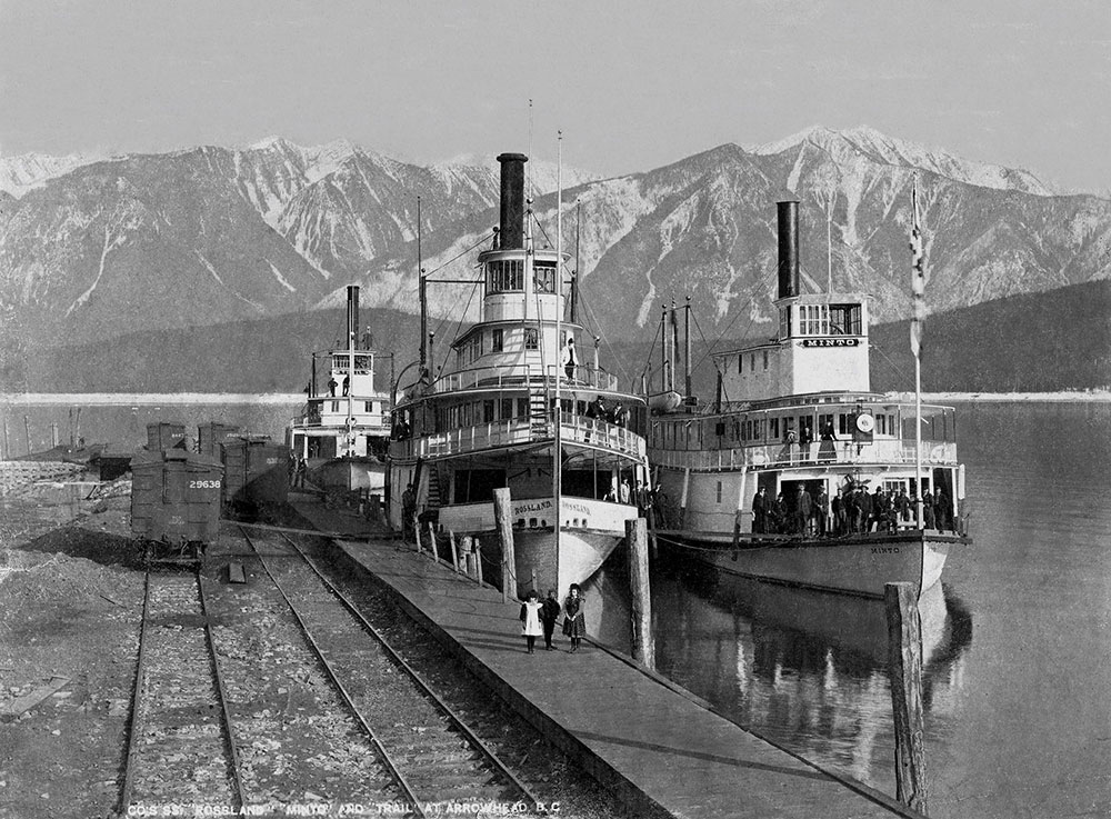 Black-and-white photograph. Three steam ships sit side by side on the water beside a dock. People are on the ships, and a few stand on the dock. Railway lines are beside the dock. Mountains are in the background.