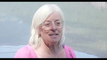 Eileen Pearkes - a white woman with white hair. She is wearing white glasses, a silver necklace, and a pink shirt. She is standing in front of a body of water.