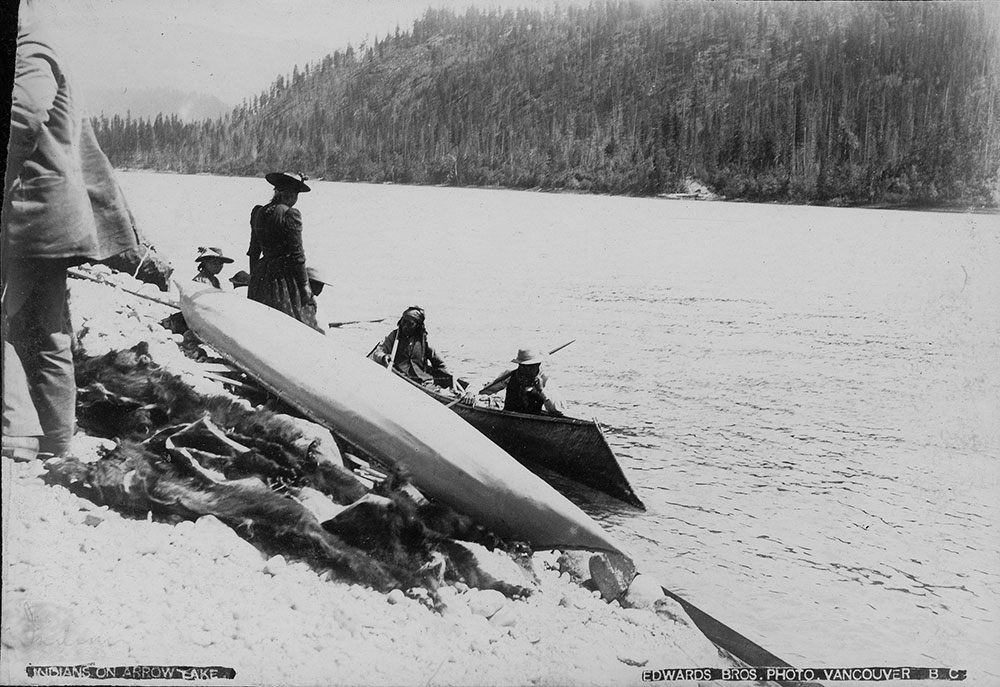 Black-and-white photograph of two Indigenous men in a canoe on the water. People in European dress are standing on the shore of the Arrow Lakes. Pelts are piled up under another canoe on the shore.