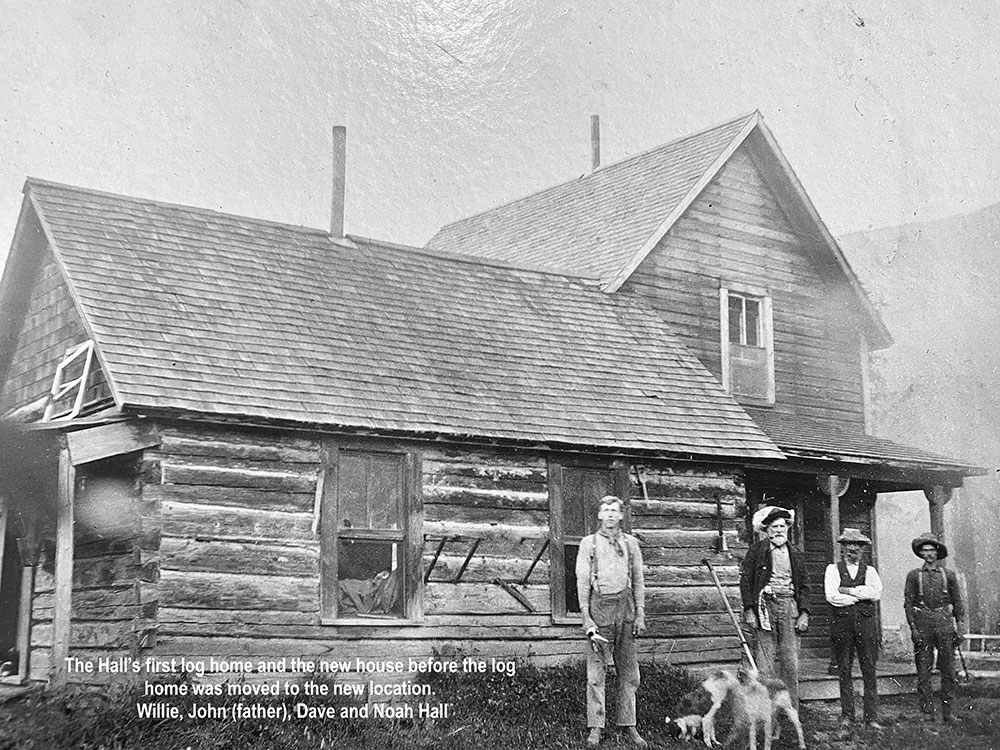 Photographie en noir et blanc montrant quatre hommes et deux chiens devant une maison en bois rond. Une légende apparait au bas de la photo, à gauche.