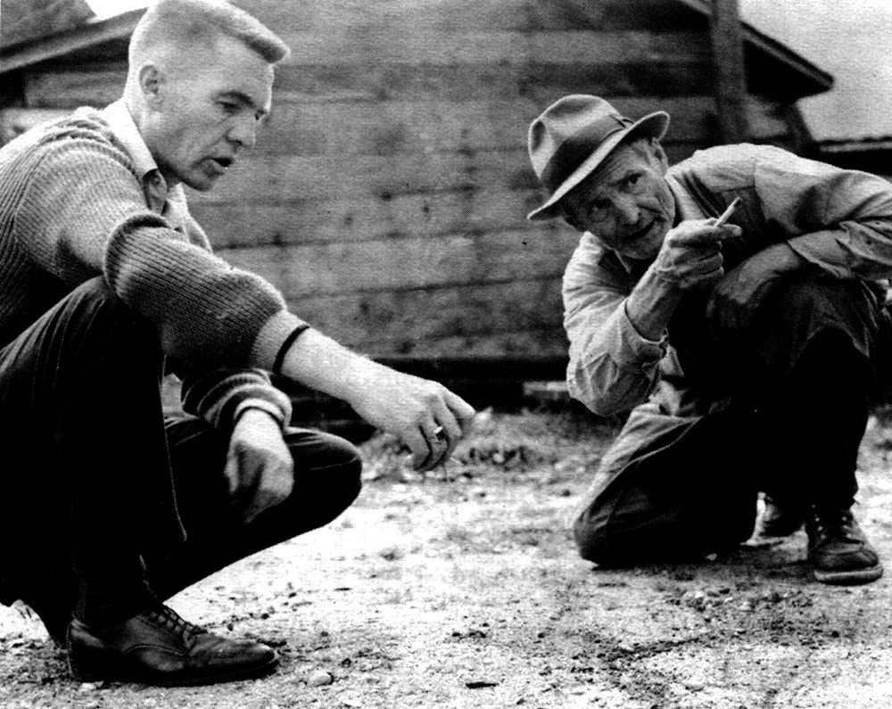A close-up black-and-white photograph of two men crouching near the ground with serious expressions. The man on the right is pointing with his pen. Part of a log building can be seen in the background.