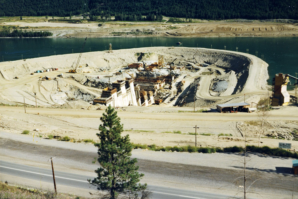 A colour photograph showing a dam under construction. A paved road is in the foreground, with a lone fir tree in the middle.