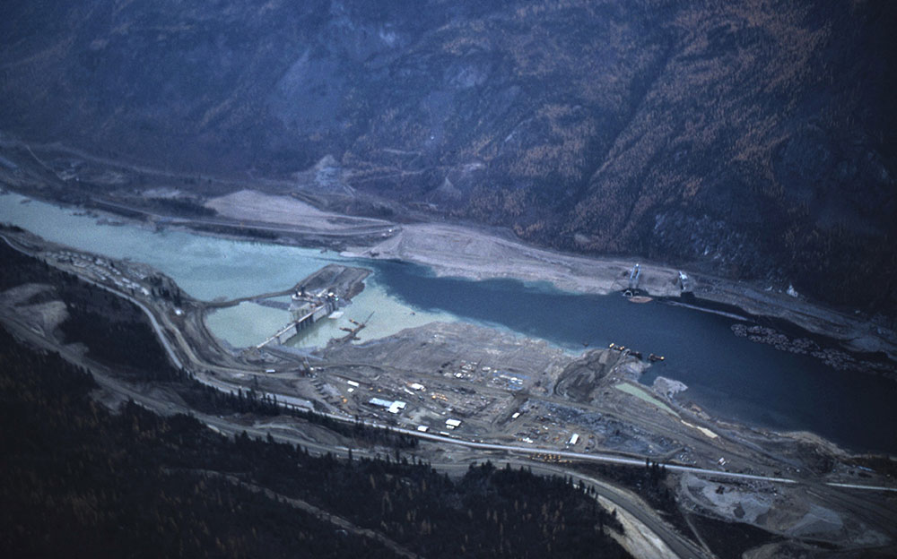 A birds-eye-view panoramic photograph of the dam under construction. The image shows the construction site, the dam, the water, and the mountains on the other side.