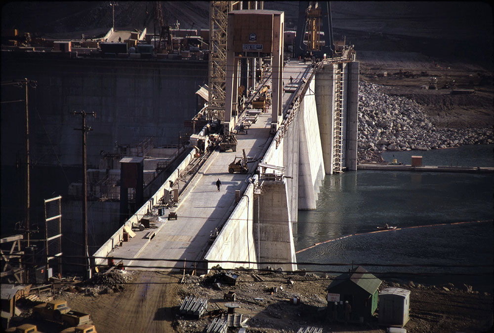 A close-up photograph of the dam under construction, nearly complete. People and construction vehicles are on the dam.
