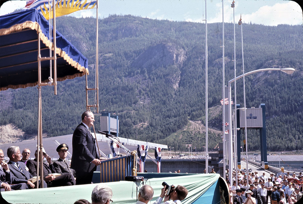 W.A.C. Bennett stands smiling at a podium. Flags and men sitting behind him. A photographer is standing in front with other men taking a photo. The dam can be seen on the right side with a crowd of people in front. Mountainside in the background.