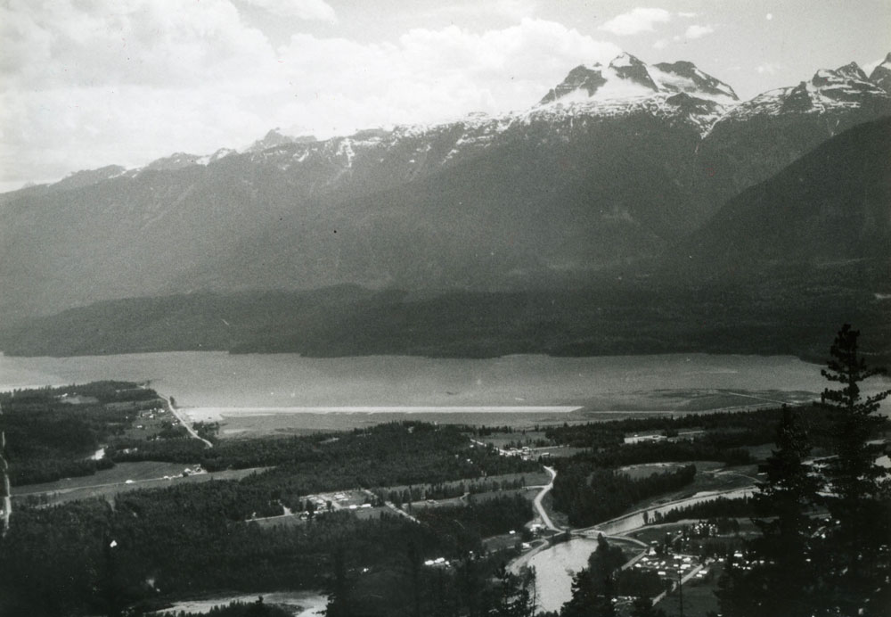 Black-and-white aerial photograph of Revelstoke that shows mountains, a river cutting across the photograph, and a section of the city. A long airstrip juts out into the water in the middle of the photograph.