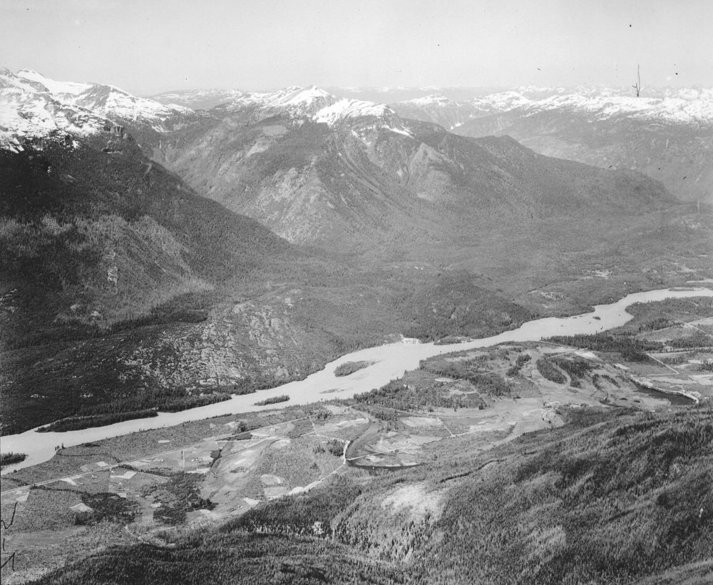 Black and white photograph showing a river bisecting the image, with high mountains on one side, and a valley with cultivated farms on the other.