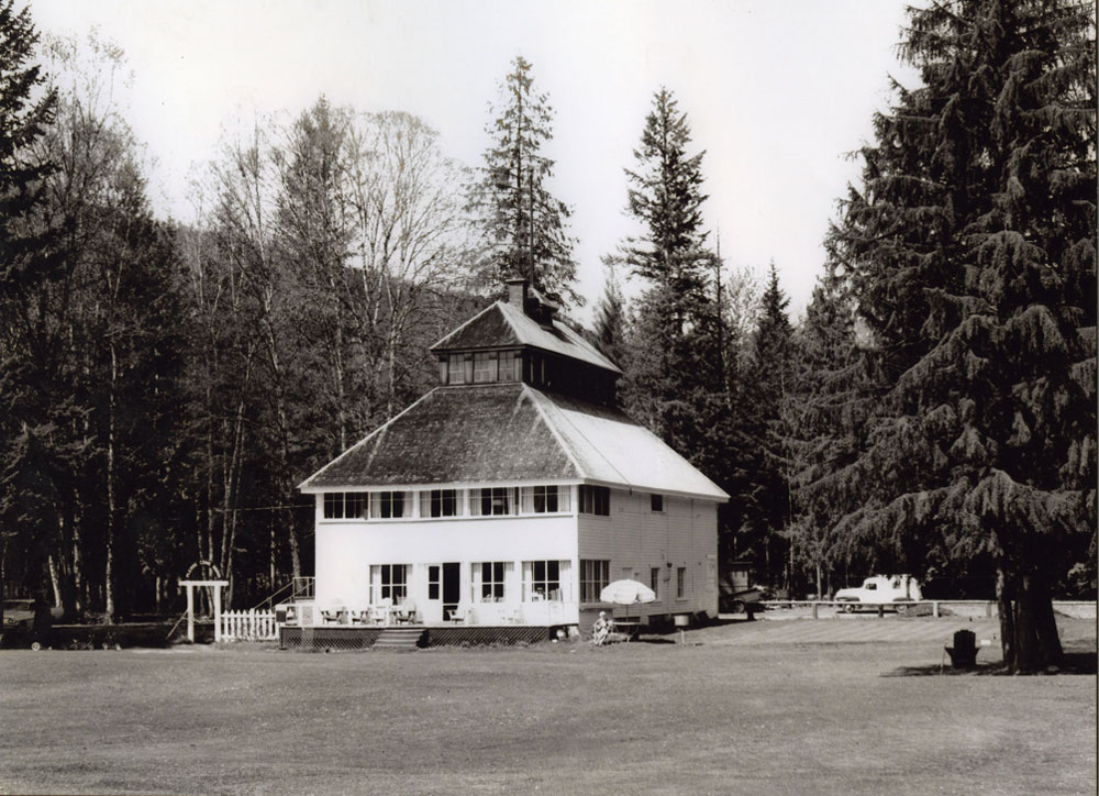A black-and-white photograph of a two-story club house sitting in a field. The building's patio has outdoor chairs on it. A women is sitting under an umbrella beside the building. There is a fence to the left of the building. Trees and a car on the road in the background.
