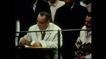 A photo of a man in a cream-coloured suit jacket signing a paper. Other people seen standing around him in the background.