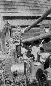 Black-and-white photograph. A boy leans against a wooden fence while holding a younger girl’s hand. Cows’ heads stick out between the fence, and they are drinking/eating from large buckets. One bucket is toppled over. Behind them are a couple of wooden buildings with shingled roofs.