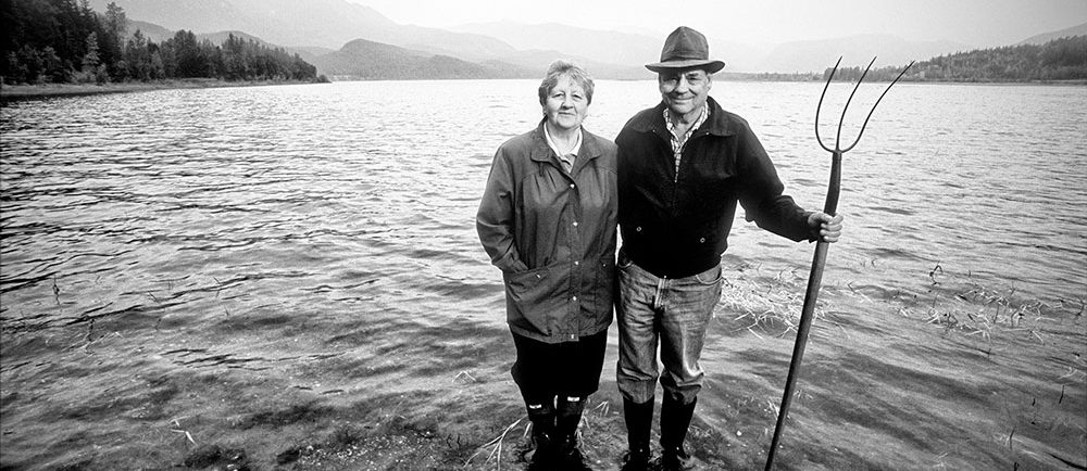 A black-and-white photograph of a woman and a man standing ankle deep in a body of water. The man is holding a pitchfork. Mountains and trees are in the background.