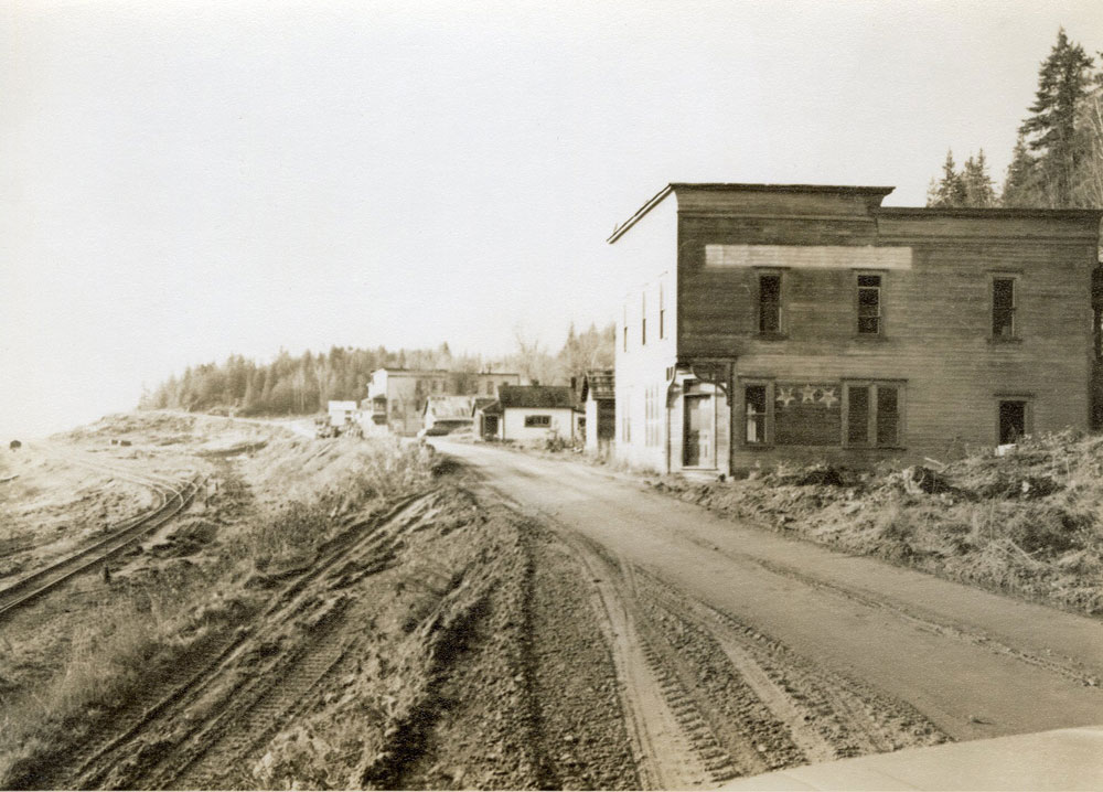 A grey-scale photo of a row of derelict buildings sitting to the right of a dirt road. Railway tracks can be seen on the left.