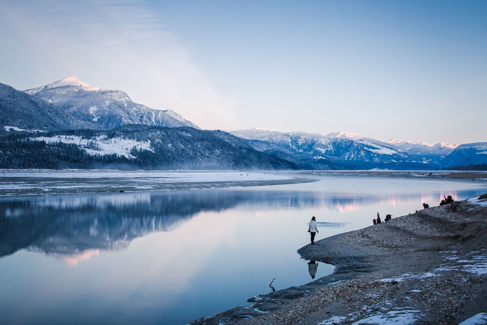 A colour photograph of snowy mountains reflecting off a body of water. A person stands on the bank beside the water.
