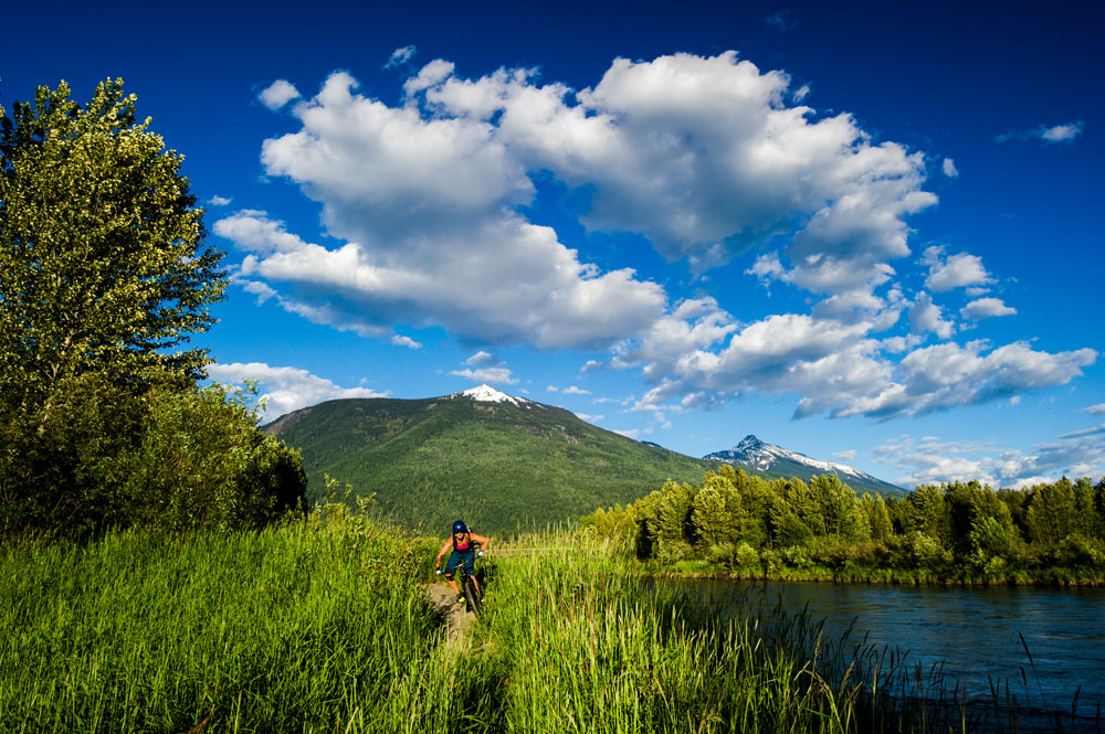 A colour photograph of a person biking down a dirt path. The path has tall greenery on either side. The river is on the right. Trees are on both sides. Green mountains with snow on the very top of the peaks are in the background. The sky is blue and partly-cloudy.