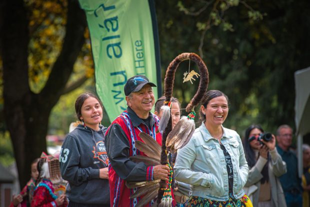 A colour photograph of an Indigenous man wearing a black ballcap with a blue feather pattern on front. He is wearing a dark blue button-down shirt, with Indigenous red and blue patterns, and holding a ceremonial staff decorated with fur, feathers, and beads. To his left is a young Indigenous woman with braided hair, and wearing a dark grey patterned hoodie. On his right is an adult Indigenous woman wearing a patterned dress and a jean jacket. All of them are smiling. Behind them are a few other people who are not in focus, and a green banner. They are standing in a treed area.