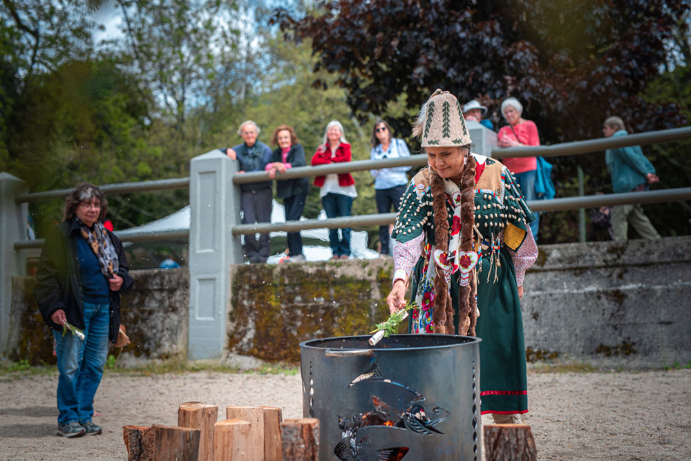 A colour photograph of a Sinixt woman, dressed in traditional attire, throwing a rolled document into a specially built fire barrel. A person, holding a similar document, stands to the left. Blurred people observe behind a raised railing in the background. Trees in the background.