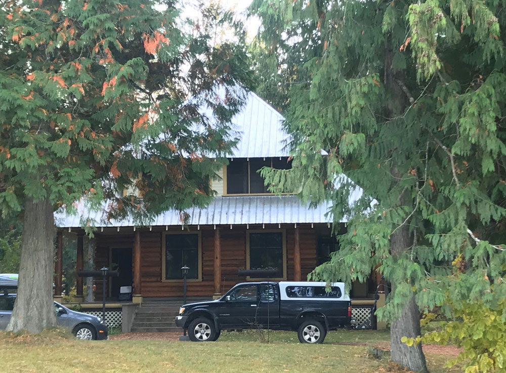 A colour photograph of a two-story log house with two large trees in front. A car and truck sit in front of the house.